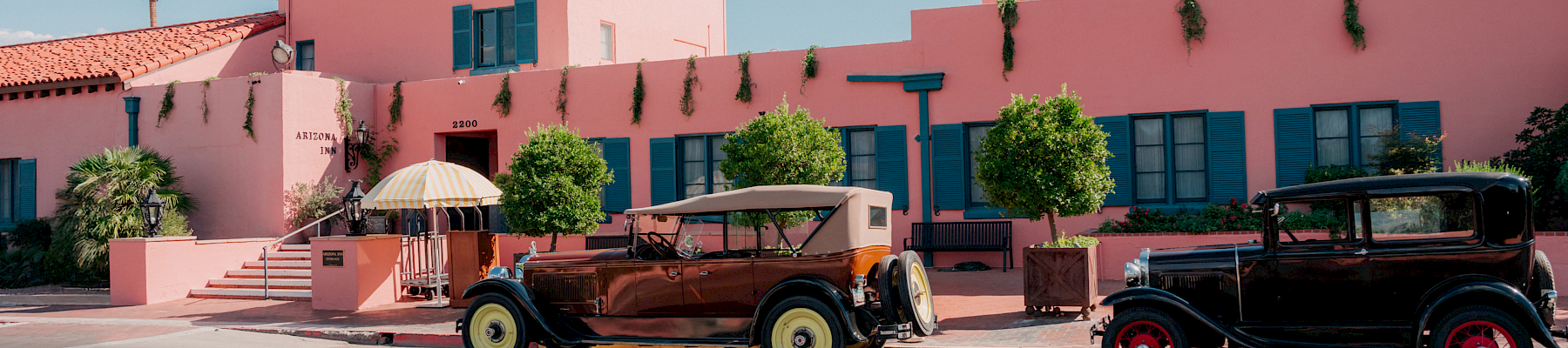 A pink resort building with blue shutters and hanging vines, plus classic cars parked in front under a striped umbrella.