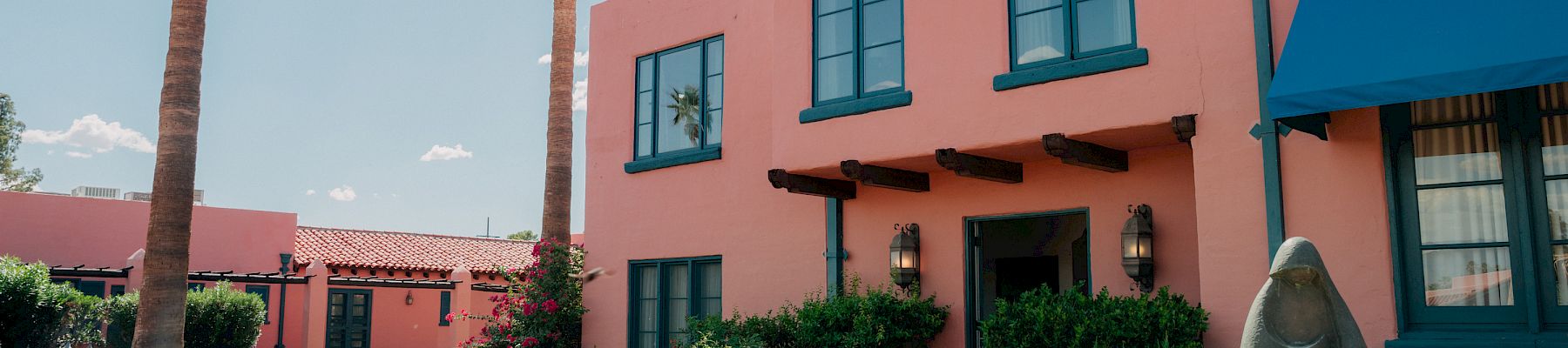 A sunny garden courtyard with blooming flowers, tall palm trees, and a pink building with blue-trim windows and a stone statue in front.