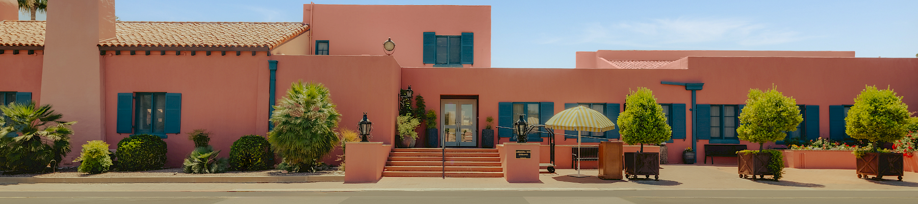 A sunny courtyard with pink stucco buildings, blue shutters, potted plants, steps, and a yellow-striped patio umbrella under a clear sky.