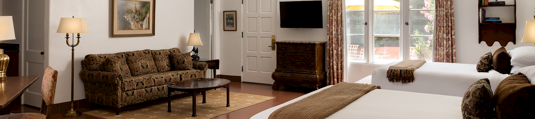 A bright bedroom with a large bed, patterned curtains, a wall TV, and a sofa in the lounge area.