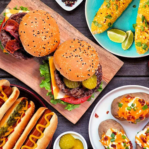 The image shows burgers and hot dogs with sides like corn, pickles, cheese, and lime wedges on plates.