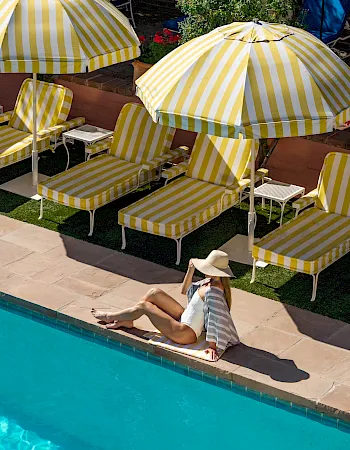 Sunlit backyard pool with striped lounge chairs and umbrellas, plus a person sunbathing by the water on the patio.