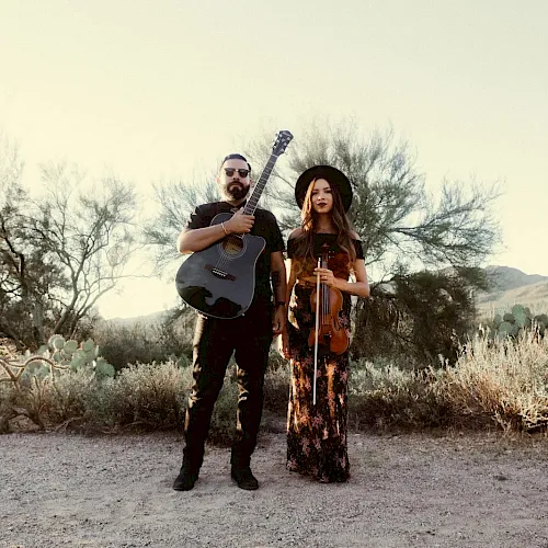 A man with a guitar and a woman with a violin stand in a desert landscape, dressed in casual, bohemian outfits, overcast sky.