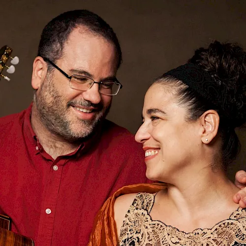 A smiling couple is posing together; the man holds an acoustic guitar while the woman looks lovingly at him.