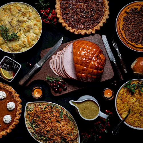 A festive meal spread with glazed ham slices, roast meat, and savory pies with bowls of sides, herbs, and cranberry garnishes.