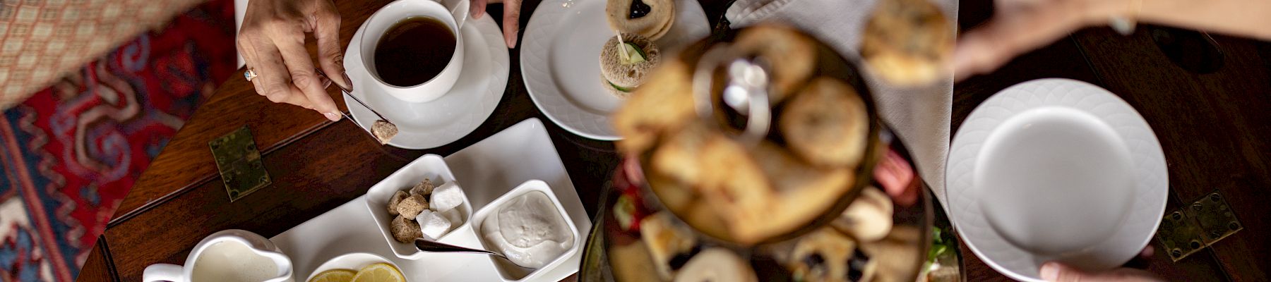 A group breakfast at a wooden table with teacups, pastries/danishes, and people serving coffee and sweets.