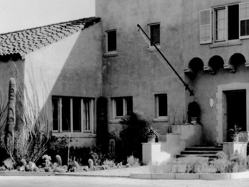 A grey stucco house with steps, arched windows, potted plants, and a small front yard on a sunny street.