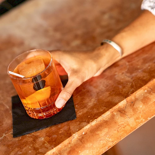 A hand rests on a bar counter with a short glass of amber liquid and a small black napkin beneath, suggesting a casual drink.