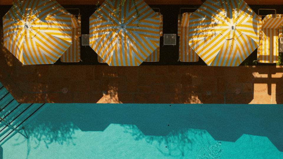 A sunny poolside scene with striped yellow-blue umbrellas, lounge chairs, and a blue water edge casting shadows over the pool.