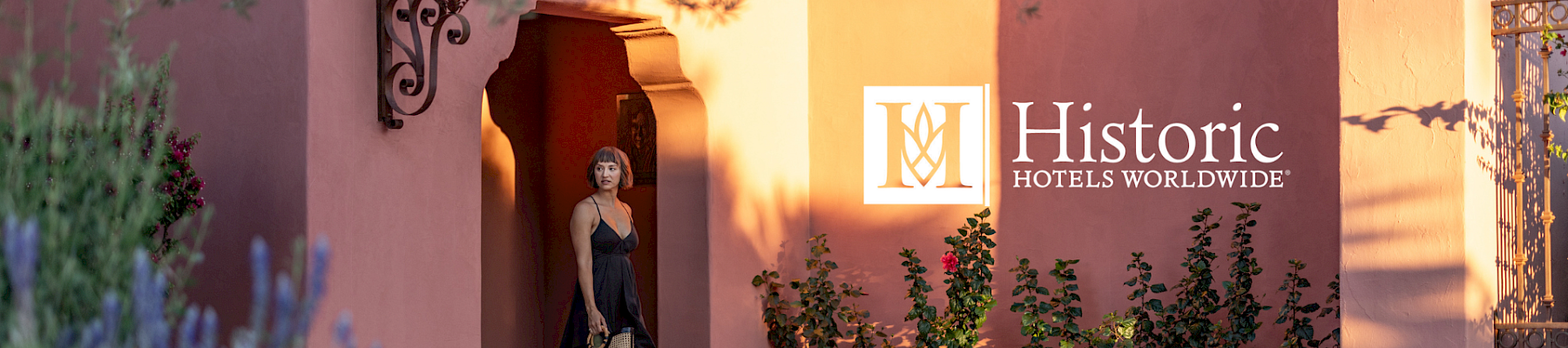 A woman stands in the doorway of a pink stucco building with plants around, beside a wall bearing the “Historic Hotels Worldwide” logo.