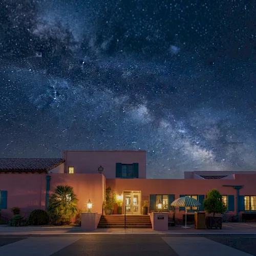 A desert-style house at night with warm lit windows under a starry sky and a visible Milky Way overhead.