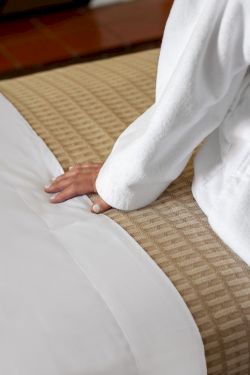 A person lies on a bed with white sheets, partially covered by a blanket, resting their hand on the mattress.