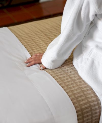 A person lies on a bed with white sheets, partially covered by a blanket, resting their hand on the mattress.
