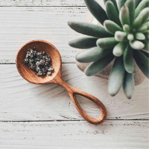 A wooden spoon filled with coarse salt sits on a white table beside a blurred succulent plant leaf.