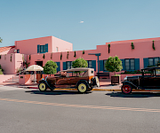 A pink resort building with blue shutters and hanging vines, plus classic cars parked in front under a striped umbrella.