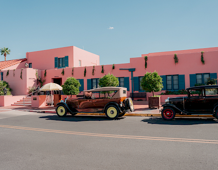A pink resort building with blue shutters and hanging vines, plus classic cars parked in front under a striped umbrella.