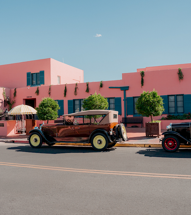 A pink resort building with blue shutters and hanging vines, plus classic cars parked in front under a striped umbrella.