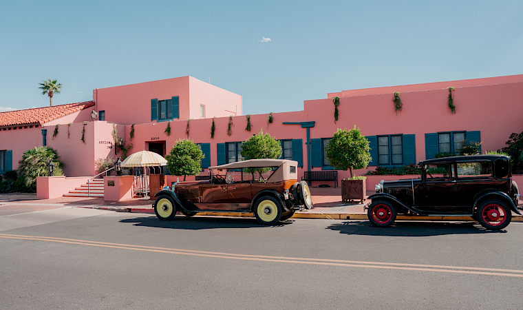 A pink resort building with blue shutters and hanging vines, plus classic cars parked in front under a striped umbrella.