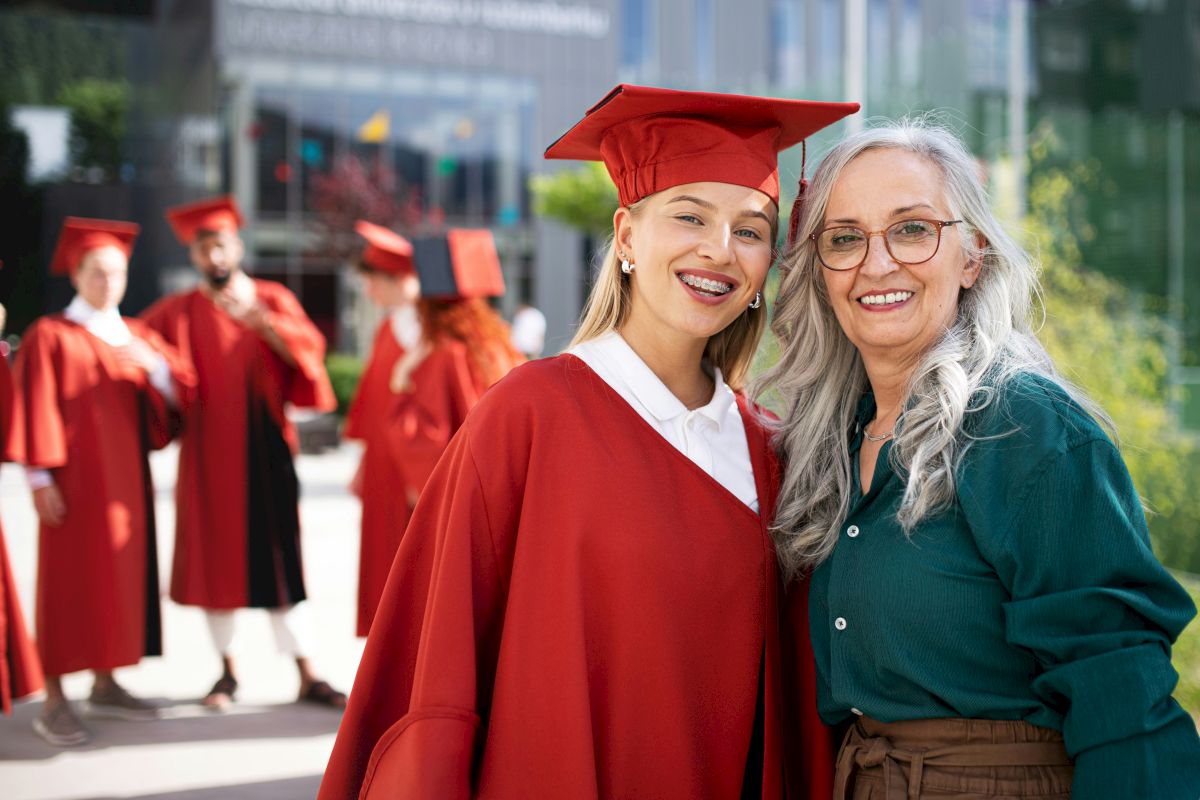 A graduate in a red cap and gown poses with an older woman outside while other graduates stand in the background.