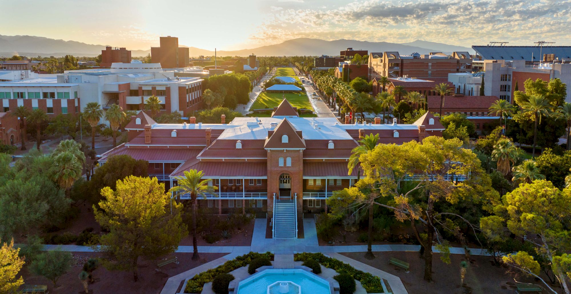 Aerial view of a campus at sunset with red-brick buildings, palm trees, a courtyard pool, and mountains in the distance.