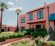 A sunny garden courtyard with blooming flowers, tall palm trees, and a pink building with blue-trim windows and a stone statue in front.