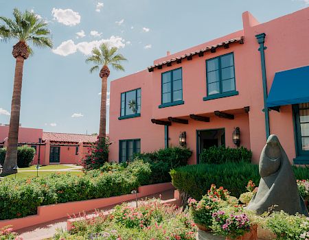 A sunny garden courtyard with blooming flowers, tall palm trees, and a pink building with blue-trim windows and a stone statue in front.