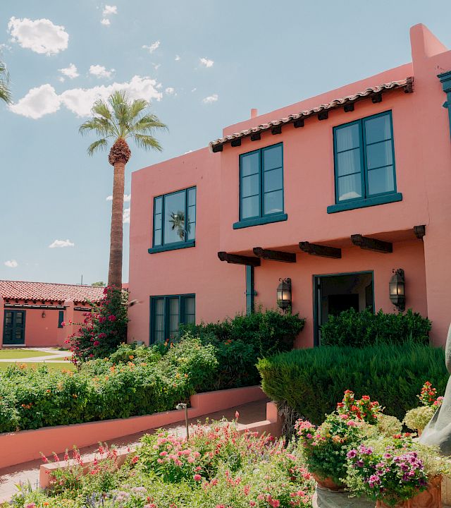 A sunny garden courtyard with blooming flowers, tall palm trees, and a pink building with blue-trim windows and a stone statue in front.
