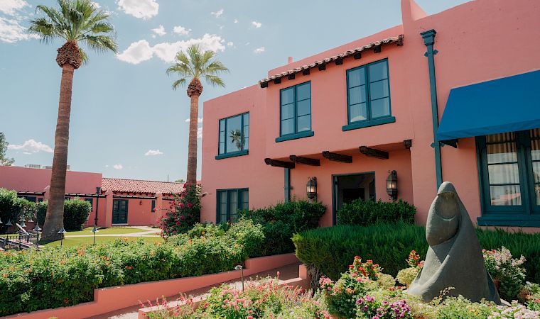 A sunny garden courtyard with blooming flowers, tall palm trees, and a pink building with blue-trim windows and a stone statue in front.