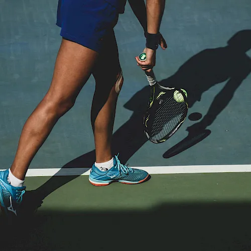 A person in blue athletic gear walks on a court holding a green paddle (likely pickleball), with a ball launcher and big shadow.