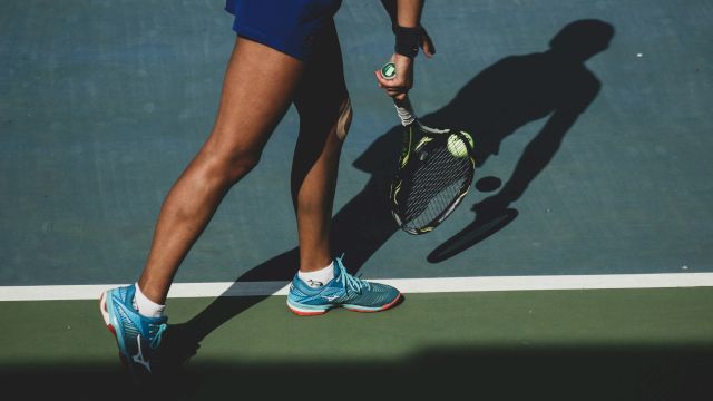 A person in blue athletic gear walks on a court holding a green paddle (likely pickleball), with a ball launcher and big shadow.