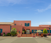 A sunny courtyard with pink stucco buildings, blue shutters, potted plants, steps, and a yellow-striped patio umbrella under a clear sky.
