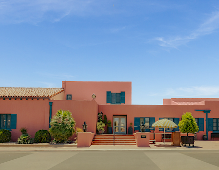 A sunny courtyard with pink stucco buildings, blue shutters, potted plants, steps, and a yellow-striped patio umbrella under a clear sky.