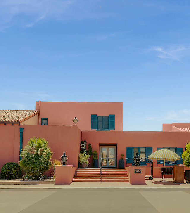 A sunny courtyard with pink stucco buildings, blue shutters, potted plants, steps, and a yellow-striped patio umbrella under a clear sky.