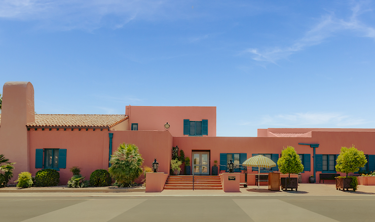 A sunny courtyard with pink stucco buildings, blue shutters, potted plants, steps, and a yellow-striped patio umbrella under a clear sky.