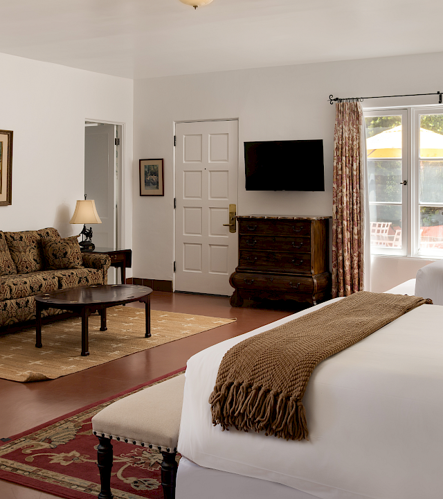 A bright bedroom with a large bed, patterned curtains, a wall TV, and a sofa in the lounge area.