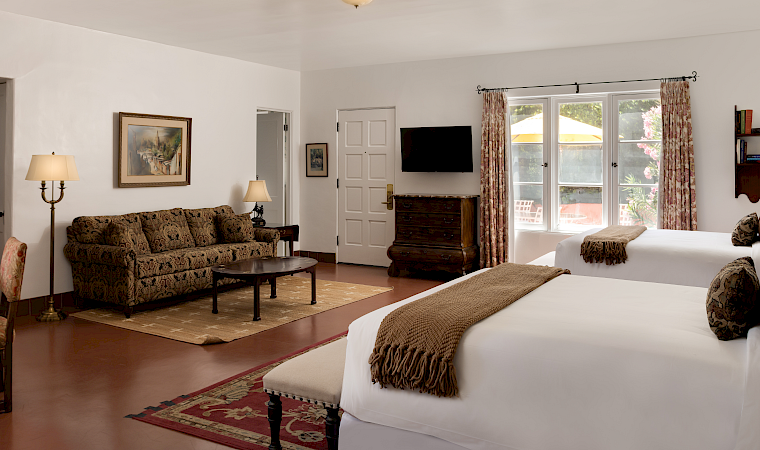 A bright bedroom with a large bed, patterned curtains, a wall TV, and a sofa in the lounge area.