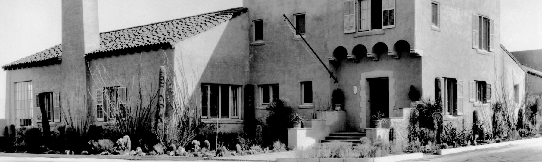 A stucco house with shuttered windows, arches, and a planted front garden, shown in a black-and-white streetscape.