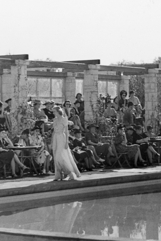 A vintage outdoor gathering: people in hats and dresses sit in rows at an event outside a brick building, with greenery around it.