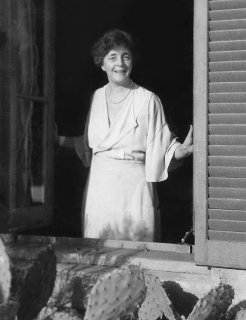 A black-and-white photo of a smiling woman in a light dress standing in a doorway, with her hand on the frame.