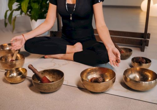 A practitioner sits on the floor, playing brass singing bowls for meditation and sound healing.