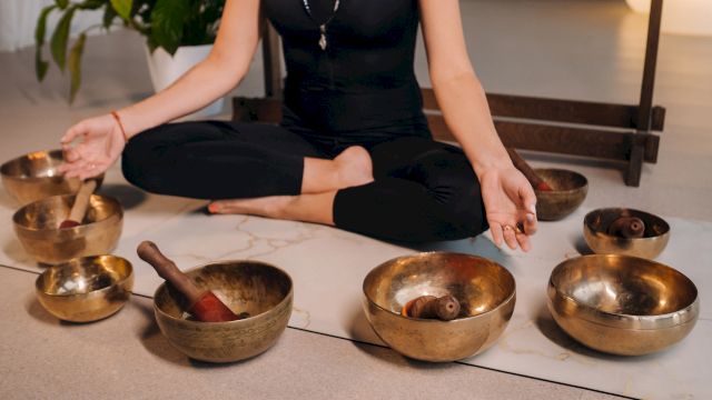A practitioner sits on the floor, playing brass singing bowls for meditation and sound healing.
