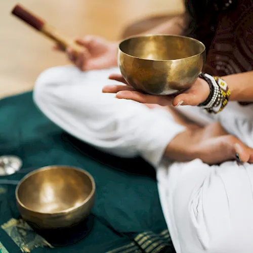 A healing scene: hands hold brass singing bowls for sound therapy, with a person preparing to play them during a calm session.