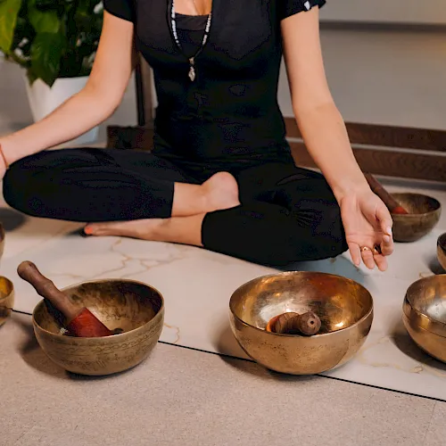 A person sits cross‑legged in a sound bath, handling brass singing bowls on a mat to make calming tones.