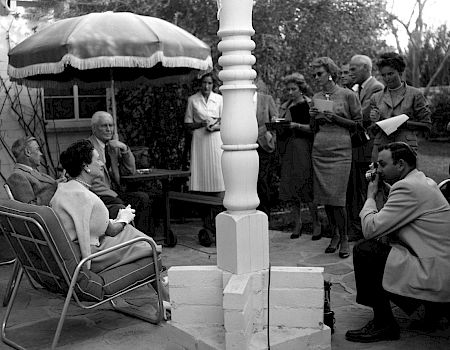 Black-and-white outdoor gathering: people sit and stand beside a gazebo while a man speaks into a microphone.