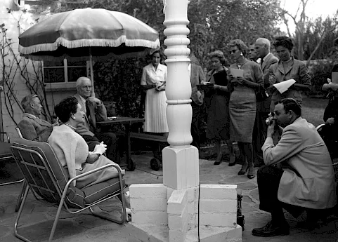 Black-and-white outdoor gathering: people sit and stand beside a gazebo while a man speaks into a microphone.