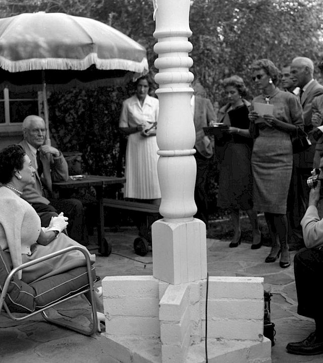 Black-and-white outdoor gathering: people sit and stand beside a gazebo while a man speaks into a microphone.