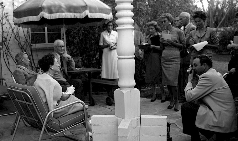 Black-and-white outdoor gathering: people sit and stand beside a gazebo while a man speaks into a microphone.