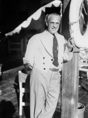 A smiling man in a white tuxedo holds a rope at a formal event, standing under a decorated canopy.