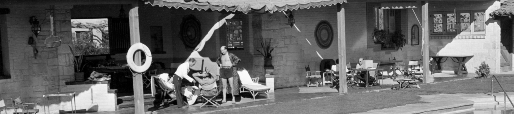 In a black-and-white courtyard, people lounge by a pool in front of a stone house with awnings and garden chairs.