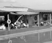 In a black-and-white courtyard, people lounge by a pool in front of a stone house with awnings and garden chairs.
